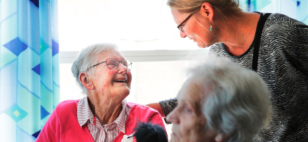 Dame der sidder med dukke kigger smilende op til dame der kigger ned på hende