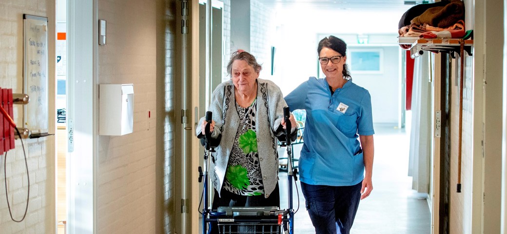 Personale der går ved siden af kvinde med høj rollator på gangen på Bakkegården